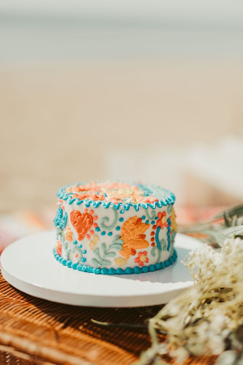Colorful floral buttercream cake with teal border on white stand, decorated with orange and yellow flowers by the beach in Port Washington