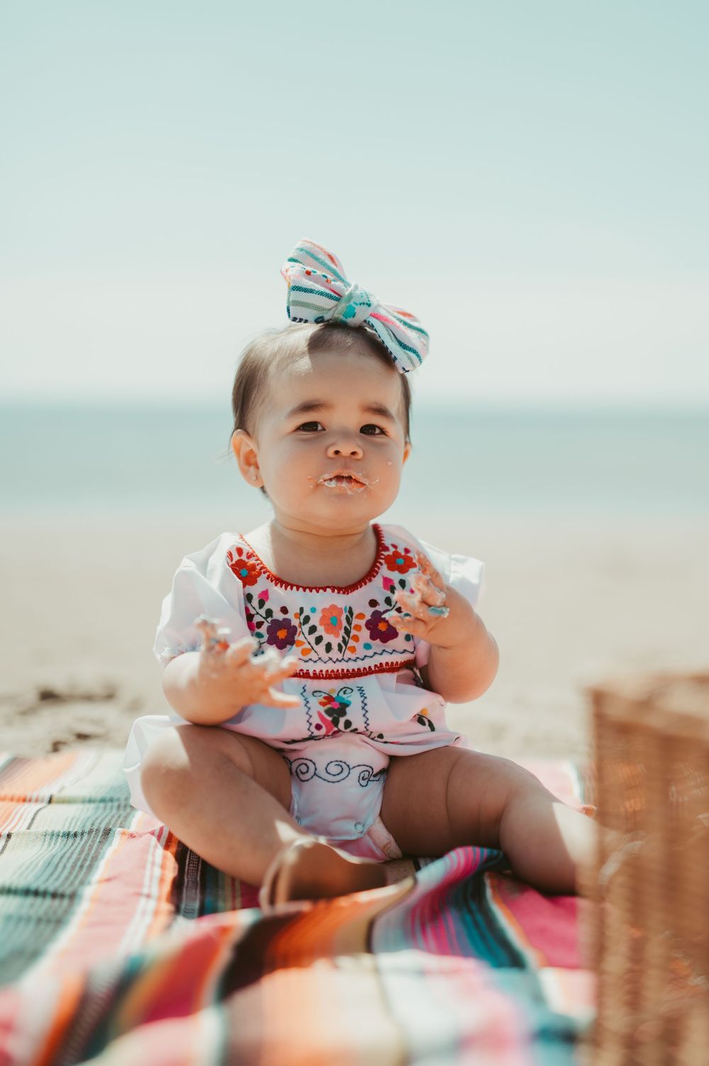 Adorable baby girl in floral embroidered dress and bow headband sitting on colorful blanket at the beach.