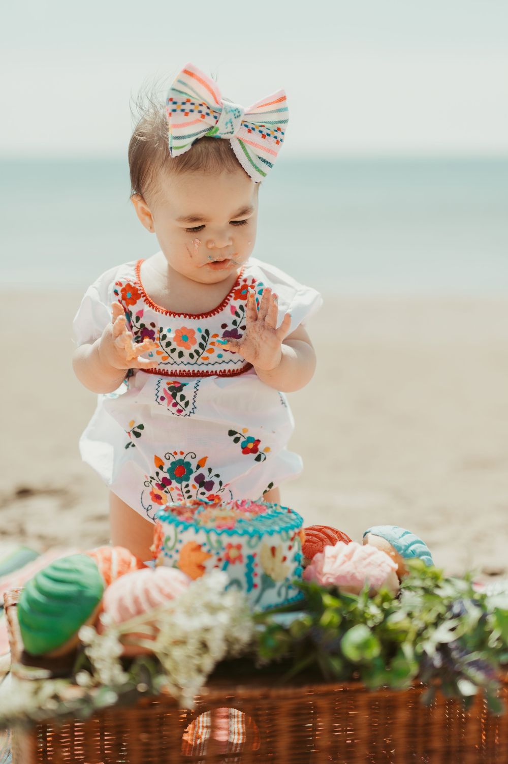 Baby girl in embroidered white outfit and bow headband plays with colorful Easter eggs on a beach.