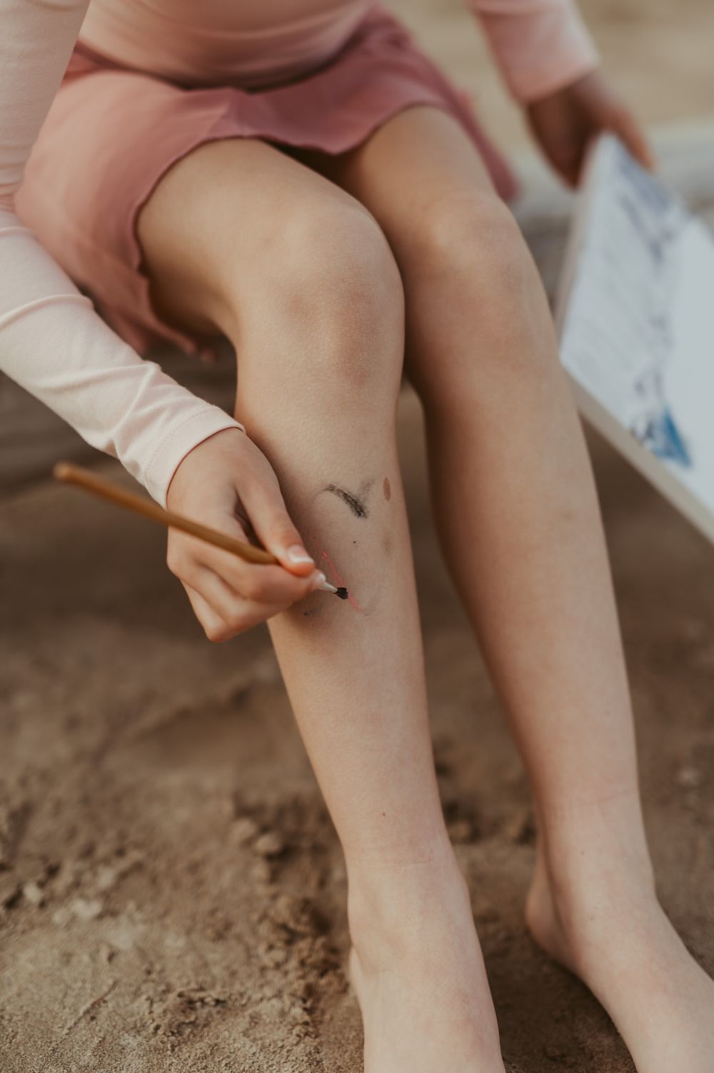 Child in pink outfit drawing on their leg with a pencil while sitting on sandy ground outdoors.