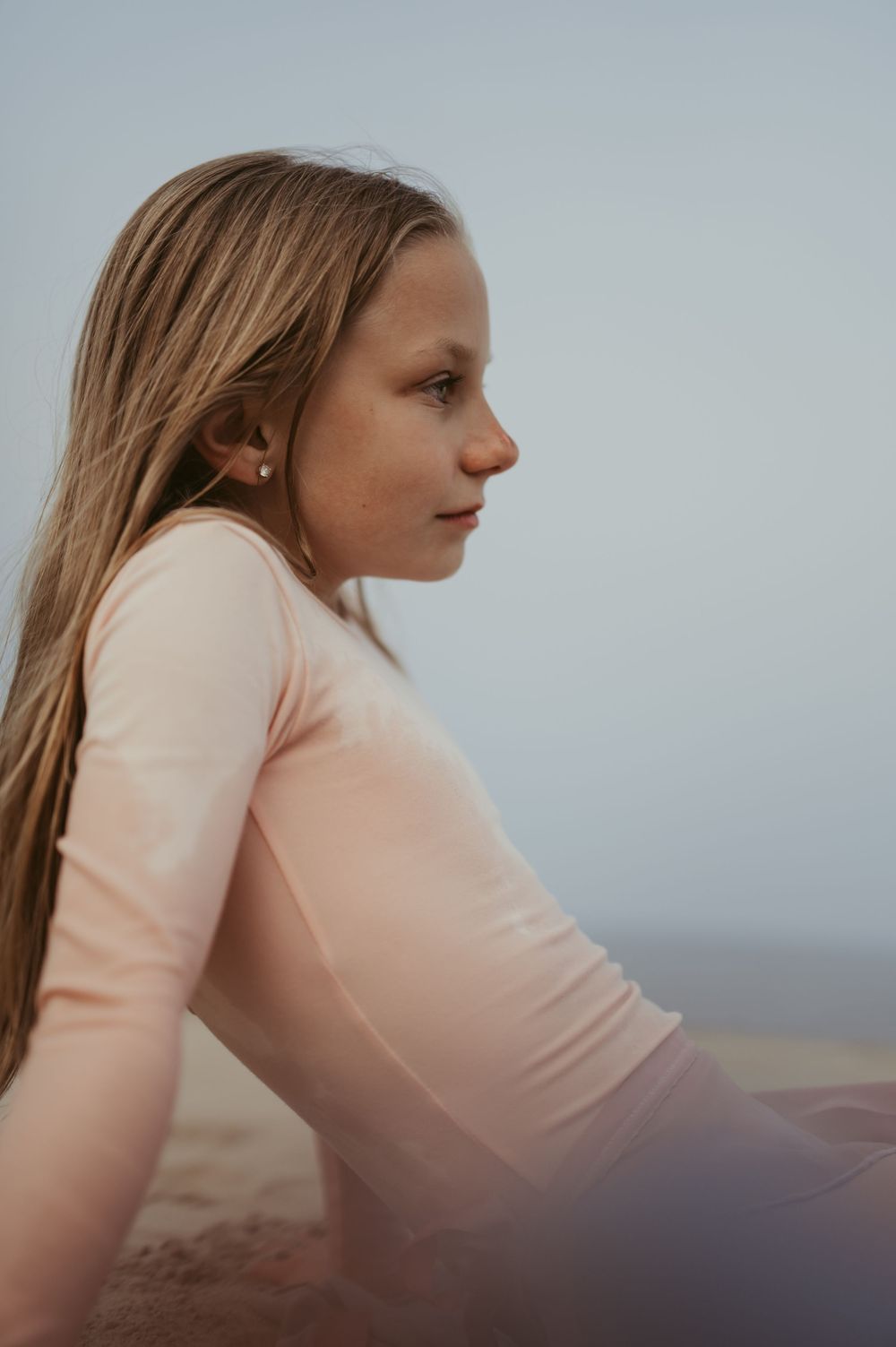 Young girl with long blonde hair sitting on a beach, wearing a pink long-sleeve top, gazing thoughtfully into the distance.