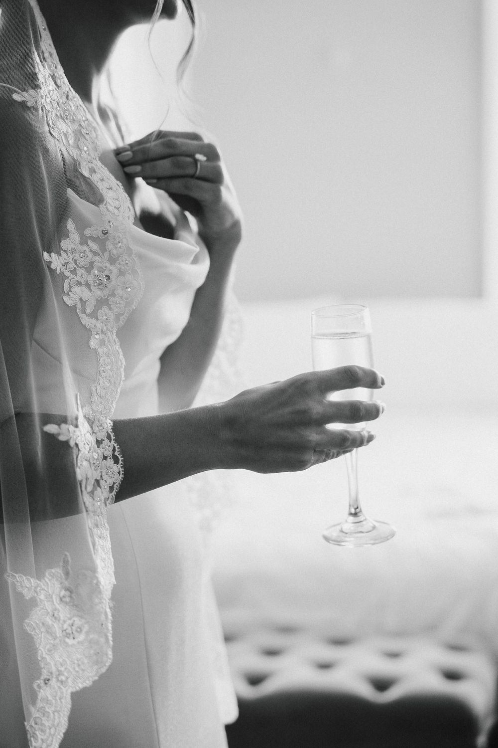 Close up of a bride in a lace veil holding a champagne glass against a light background.