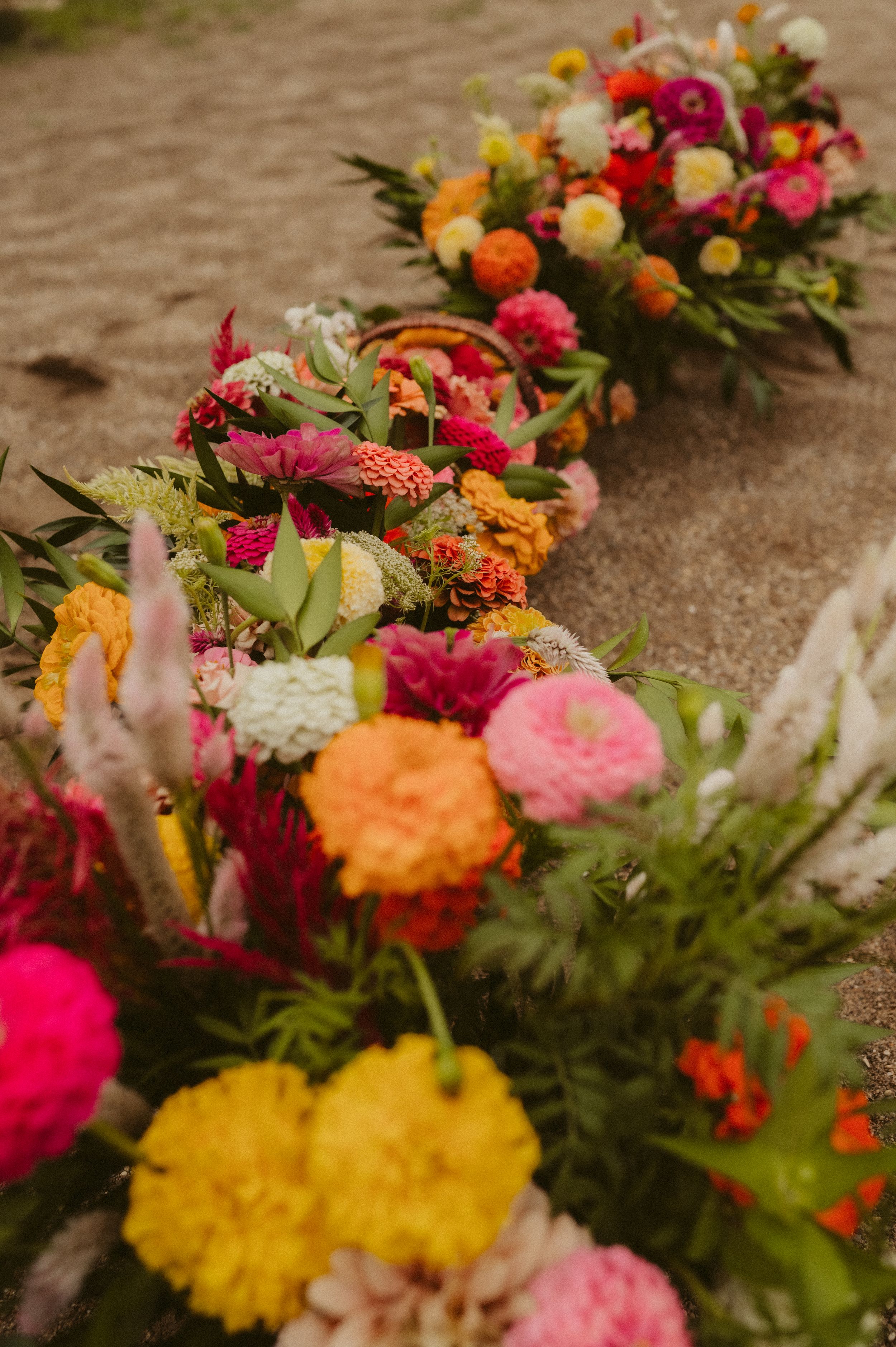 Vibrant floral arrangement with pink, yellow and orange flowers laid out in a curved line on a sandy surface.