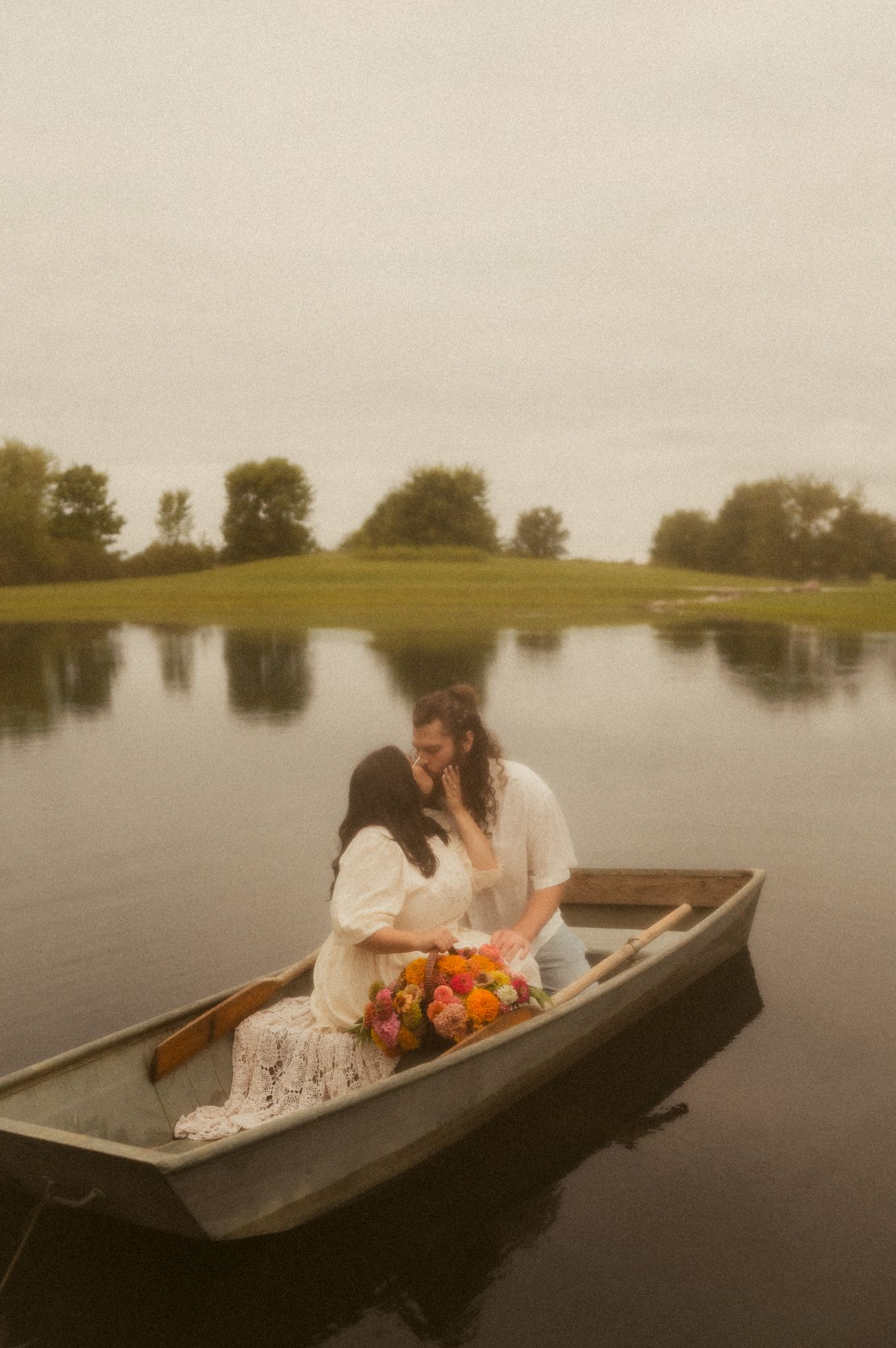 A romantic couple in white dress share intimate moments while sitting in a wooden rowboat on a misty lake.