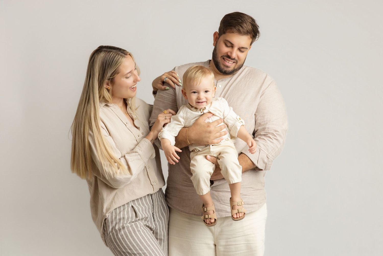 A family portrait in neutral tones features parents holding their swaddled newborn baby against a dark brown backdrop.