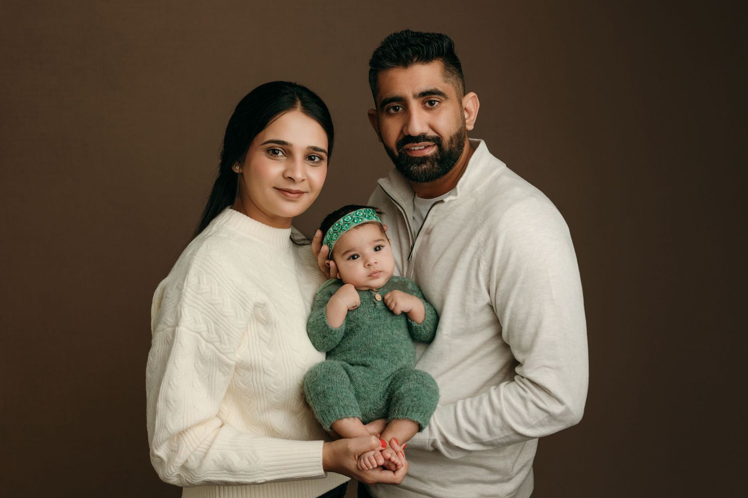 A family portrait with parents in white sweaters holding a baby wearing a green outfit taken at the studio in Victoria BC.