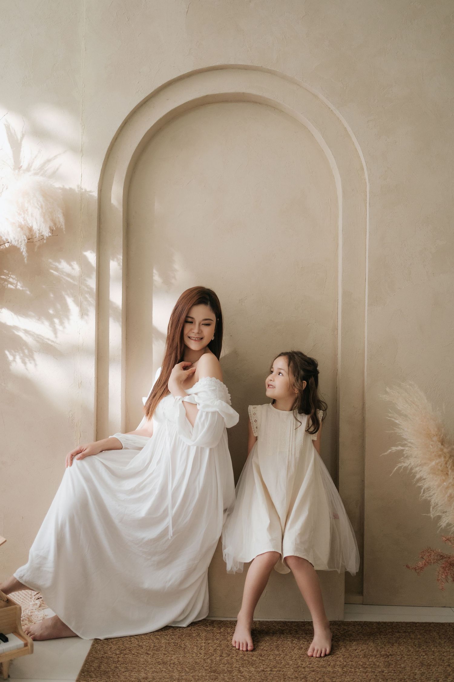 Woman and young girl in matching white dresses pose together against a beige arched wall with pampas grass decor.一位女子和一位小女孩身穿配套的白色連身裙，在飾有蘆葦草