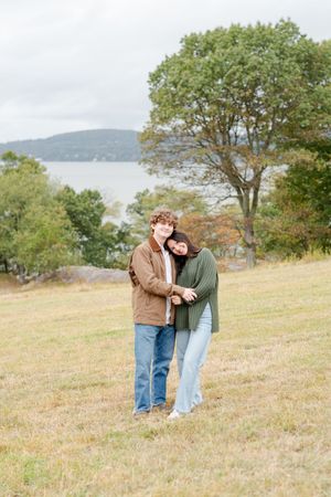 A couple stands together on a grassy hillside overlooking water and mountains on an autumn day.