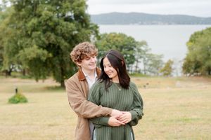 A couple embraces while walking through a scenic lakeside field with tall grass and trees in the background.
