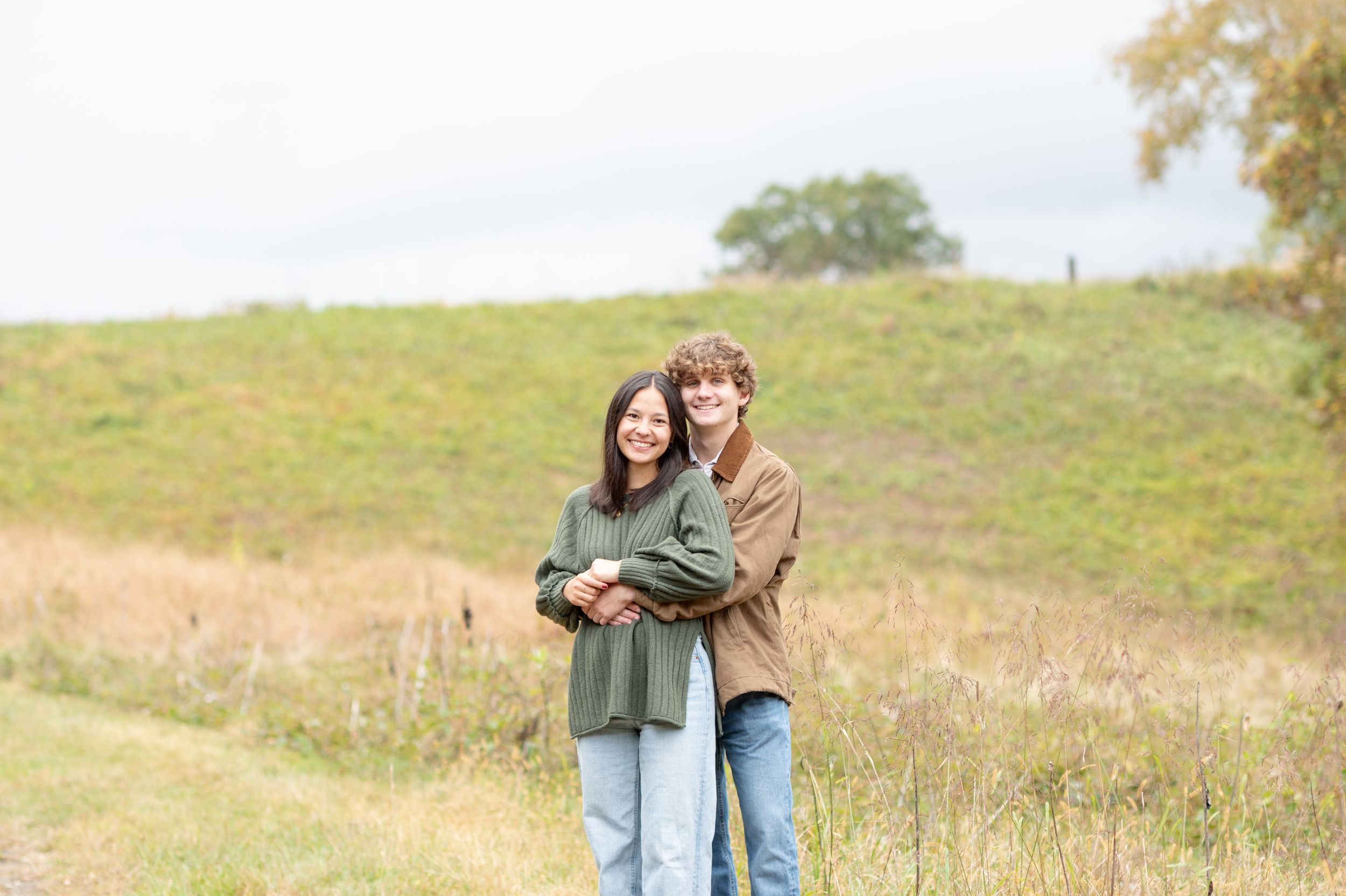 Two people embrace while standing in a sunlit meadow with golden grass and rolling hills in the background.