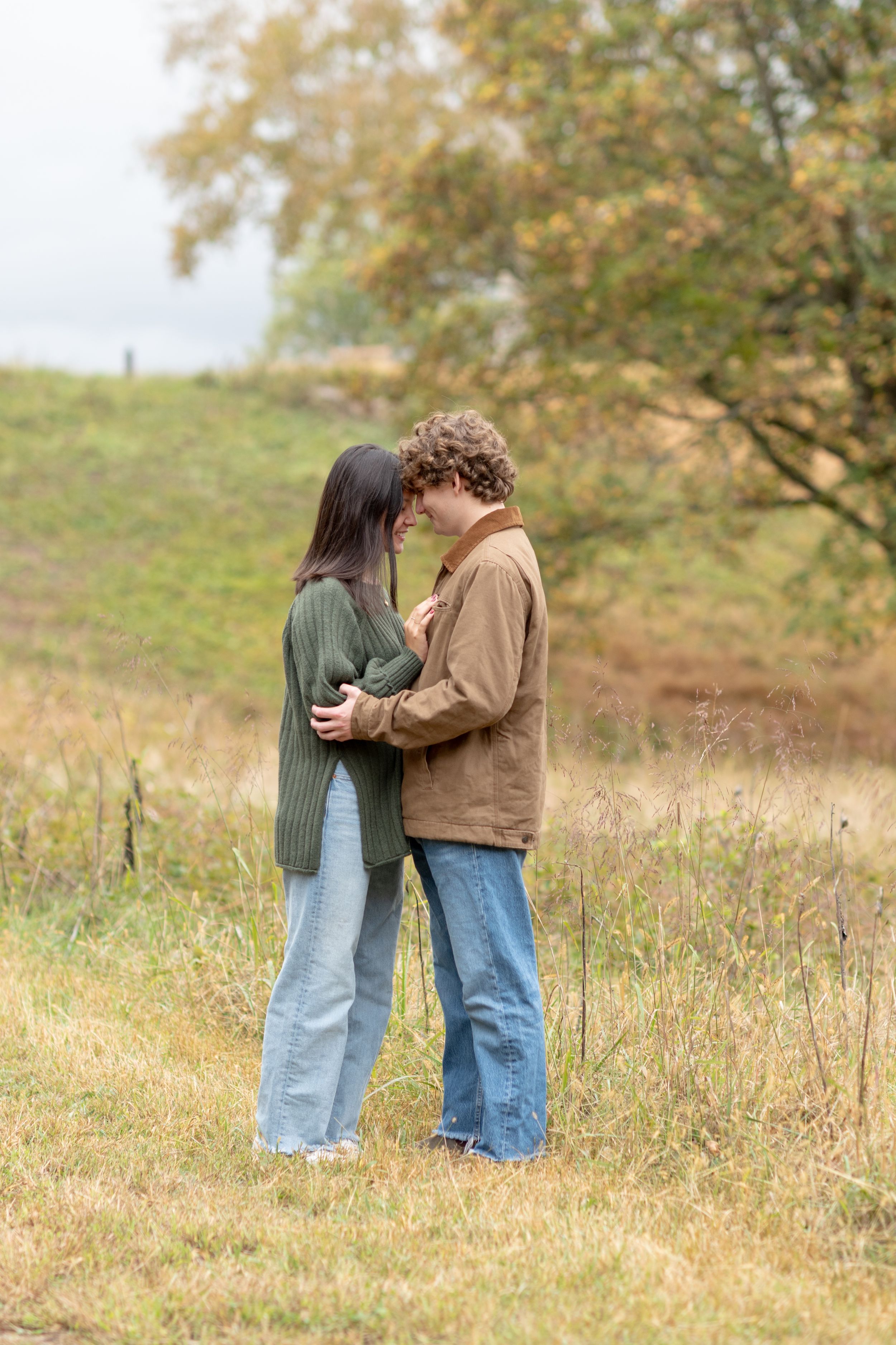 A couple stands together in a golden autumn field wearing casual denim and brown clothing.
