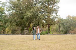 A couple stands together in a grassy field with autumn trees in the background during golden hour.