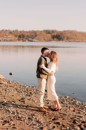 A couple shares a romantic moment on a rocky lakeshore during golden hour as autumn trees line the water's edge.