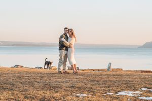 A couple stands on a scenic coastal cliff at sunset with their dog against a golden sky backdrop.