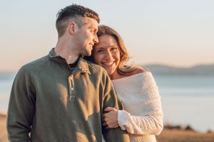 A romantic couple embraces on a beach at sunset wearing cozy sweaters in autumn.