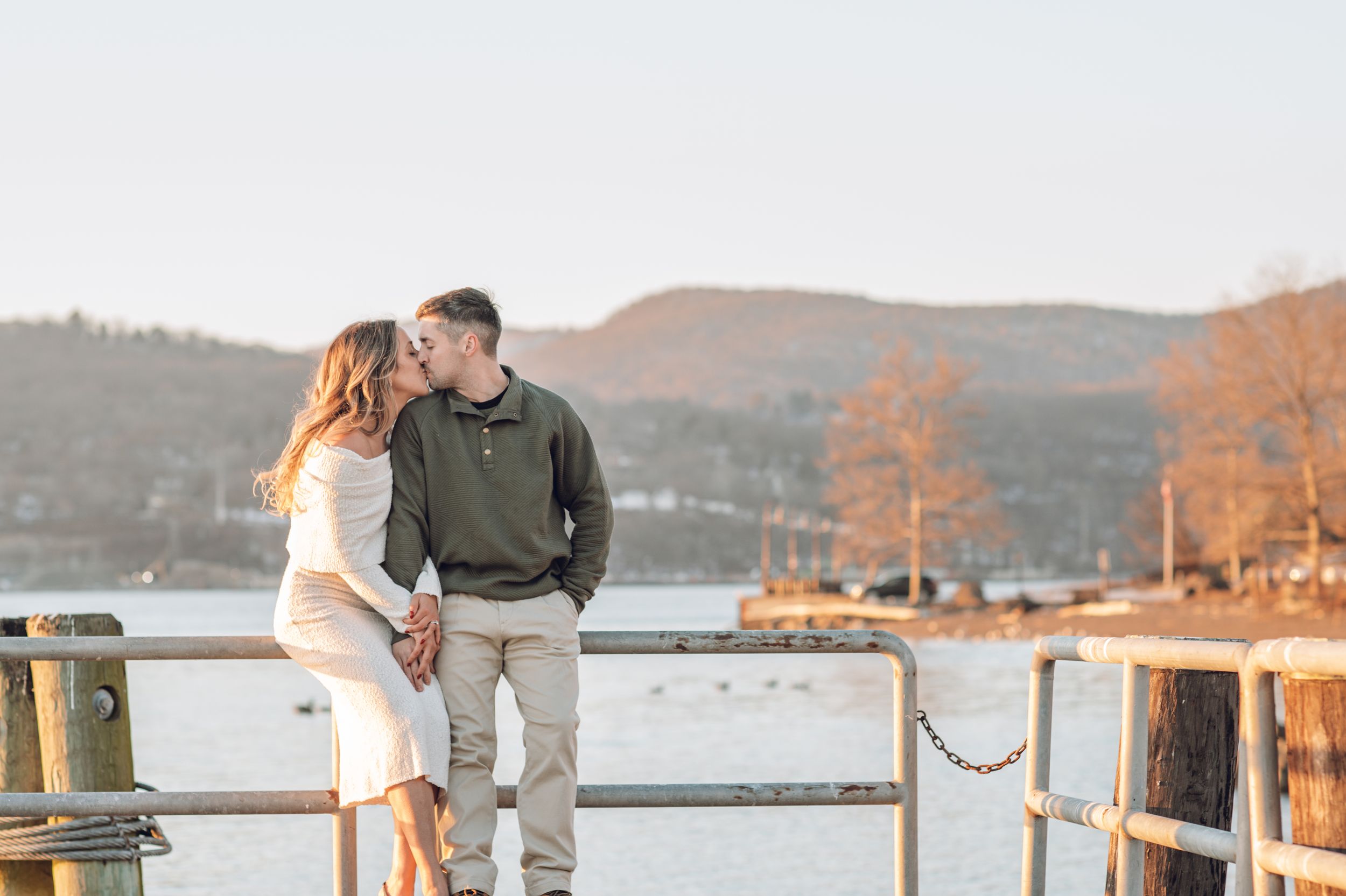 A couple shares a romantic moment on a lakeside dock during golden hour with mountains in the background.