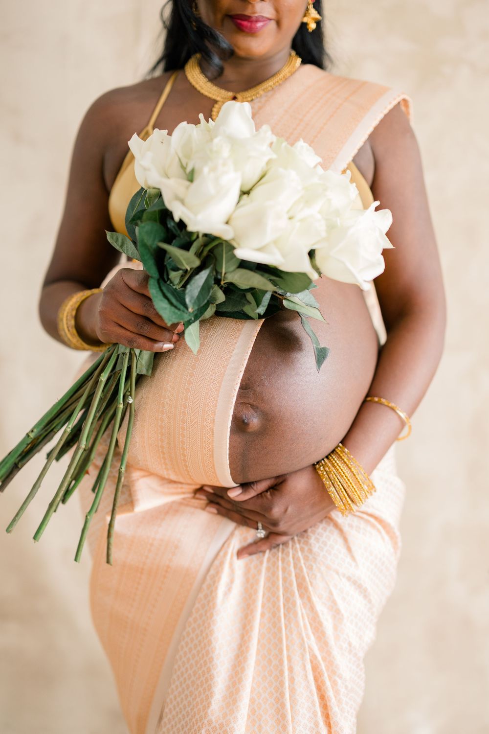 Expectant mother in peach sari holds elegant white rose bouquet during maternity photoshoot.
