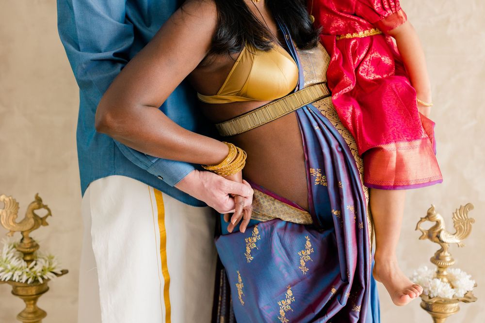 A close-up shot shows hands gently holding a baby bump while wearing a blue and red dress against a beige wall.