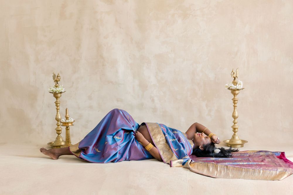 A person in colorful silk garments lies between two ornate golden candlesticks against a neutral backdrop.
