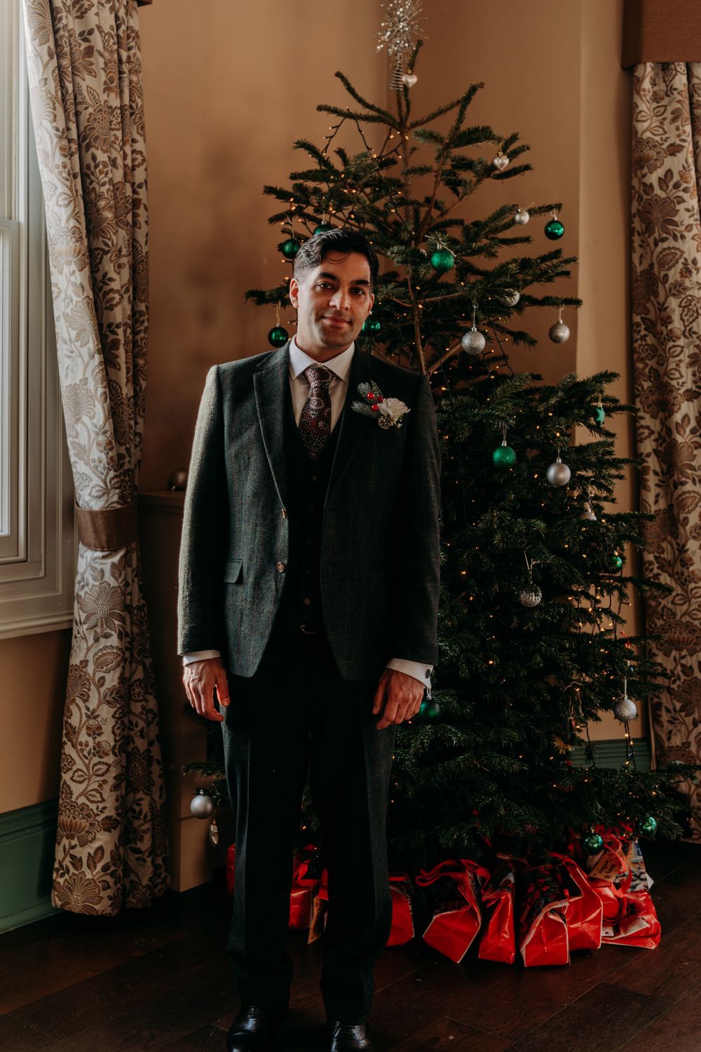 A groom in a dark suit stands next to a decorated Christmas tree with red wrapped presents underneath.
