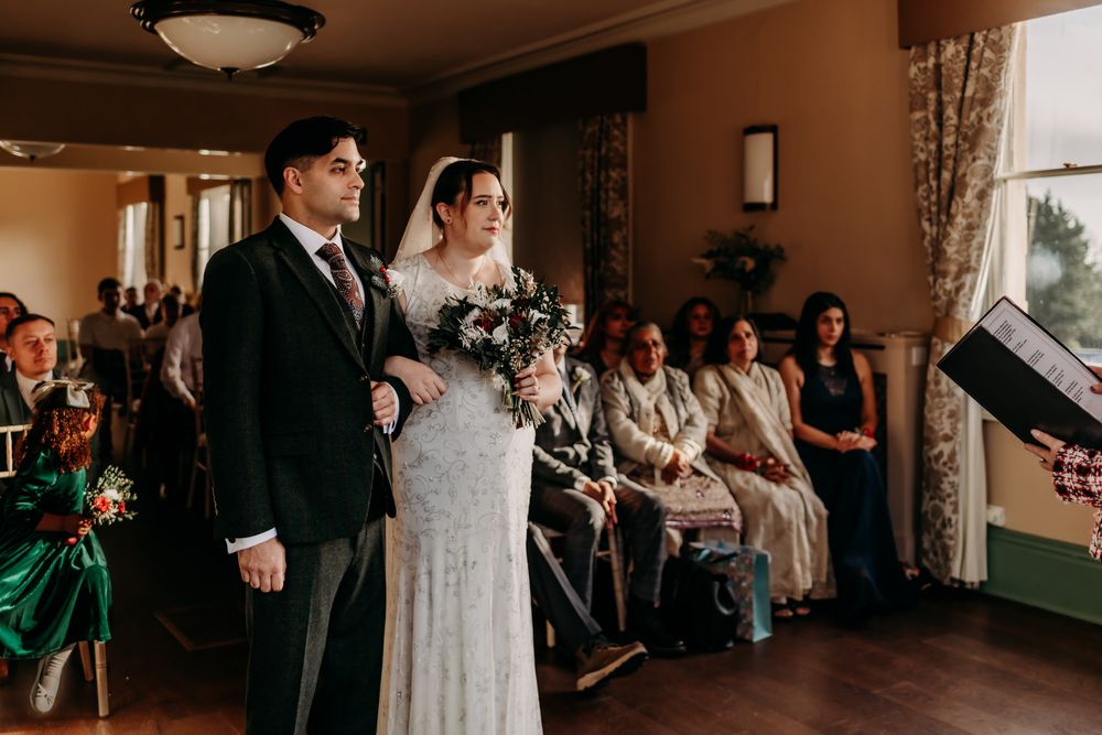 A couple stands together during a vintage-inspired wedding ceremony in a warm-lit room with seated guests.