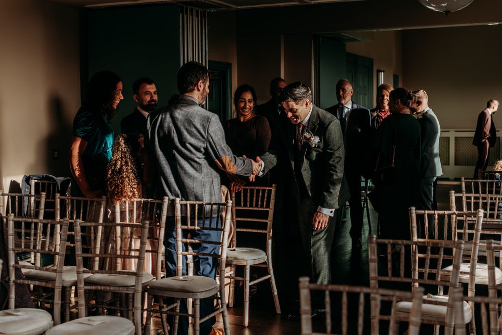 Dark silhouettes of guests standing in a dimly lit reception venue with wooden chiavari chairs.