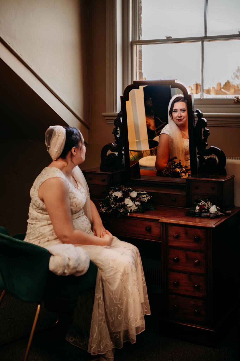 Two people in white dresses sit together in a dimly lit room with vintage furniture and window light.