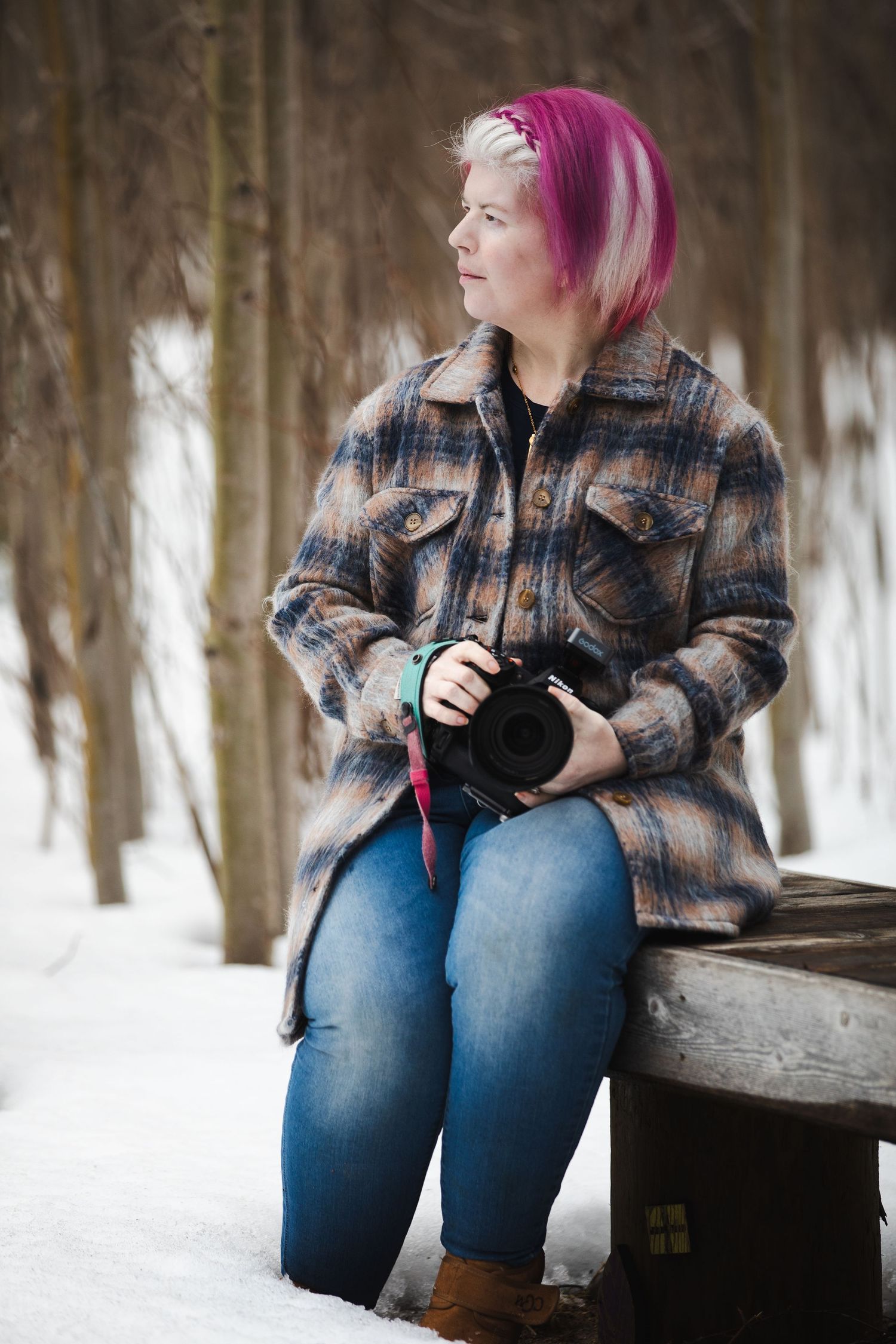 Person in plaid jacket and jeans sits on snowy log in winter forest with camera.