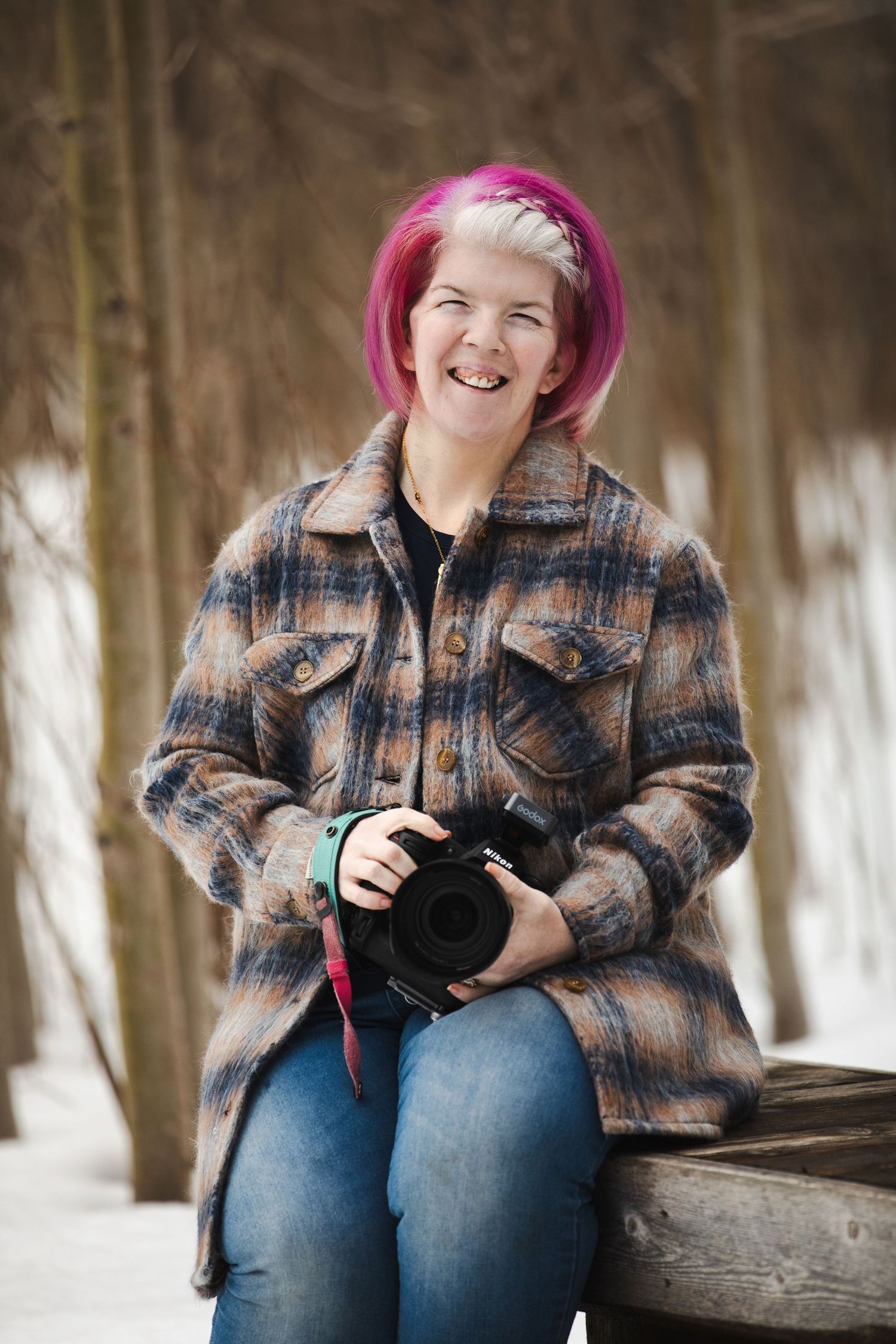 A photographer with bright pink hair wearing a plaid jacket sits on a wooden bench in a snowy winter setting holding a camera.
