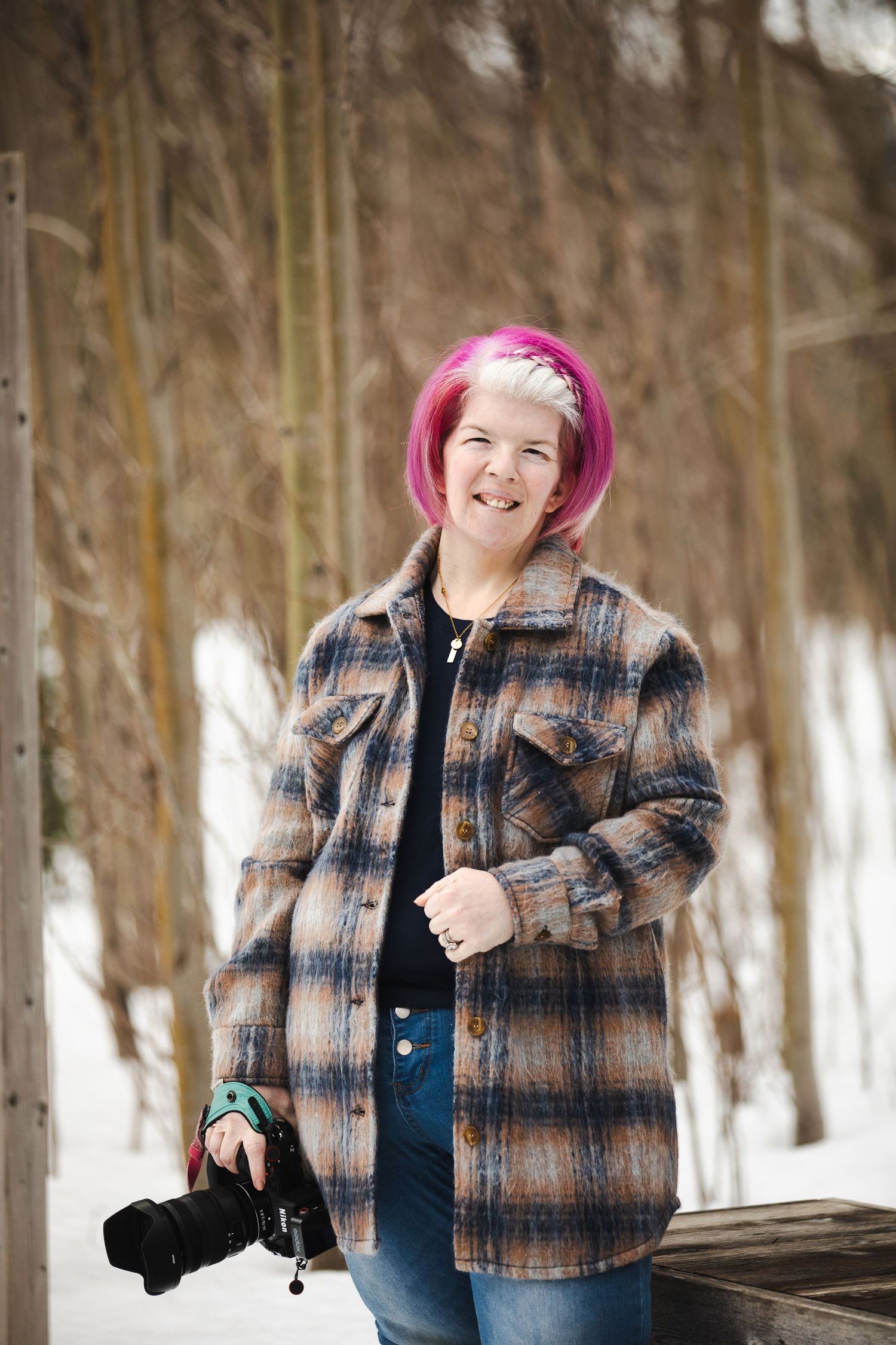 Person with bright pink hair wearing a plaid jacket poses playfully in a snowy winter forest setting.