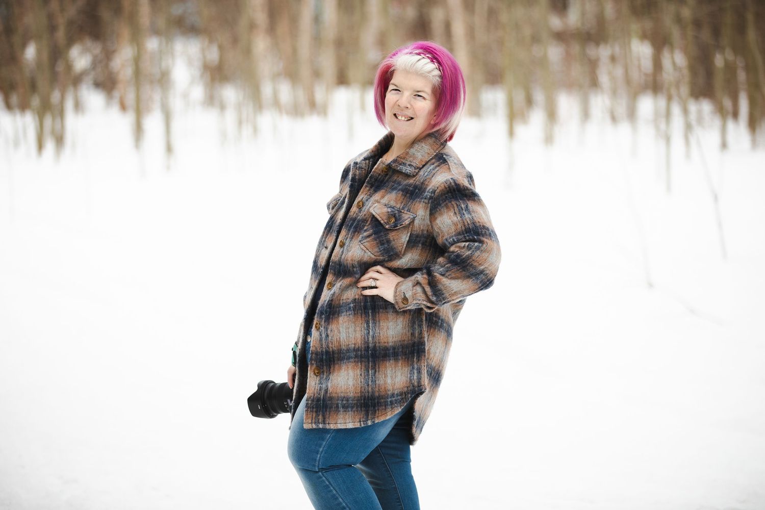 Person with pink hair wearing plaid jacket poses in snowy winter woods setting.
