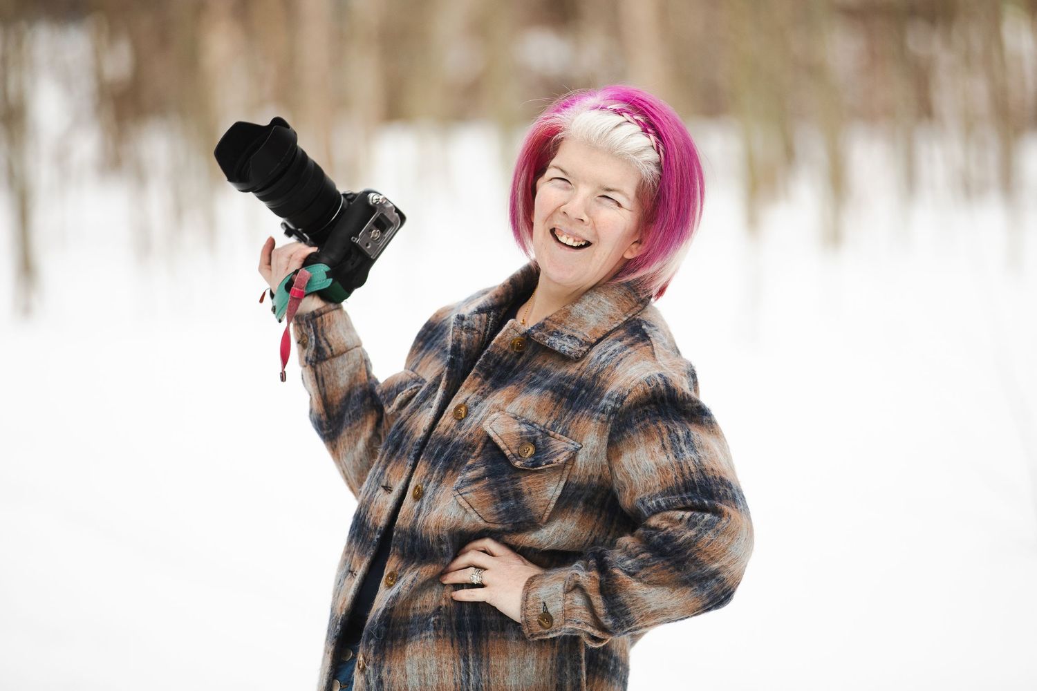 A person wearing a plaid coat and pink hair poses playfully with a camera in a snowy winter setting.