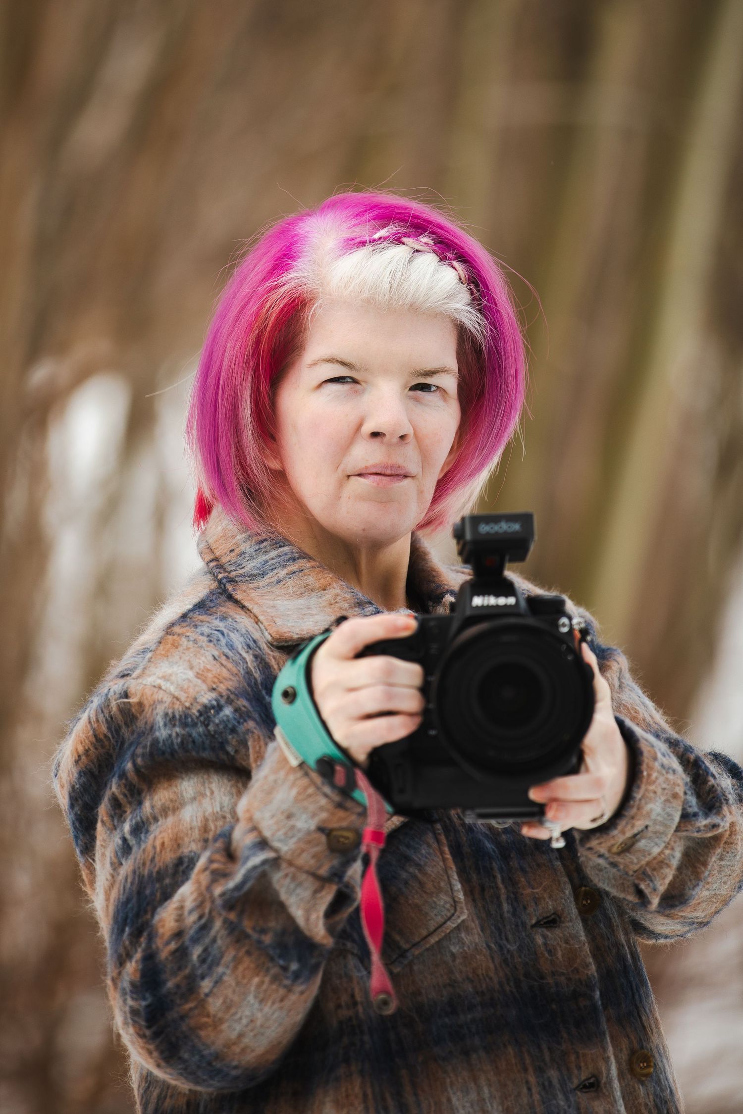 Photographer with bright pink hair wearing a brown jacket holds a camera in a wooded outdoor setting.