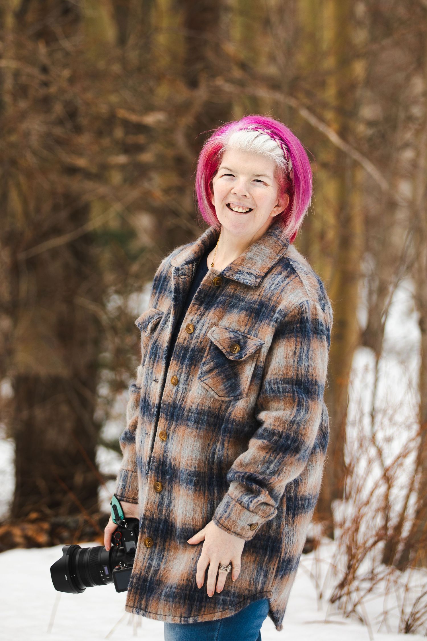 Person with pink hair wearing a plaid jacket poses in a snowy winter landscape holding a camera.