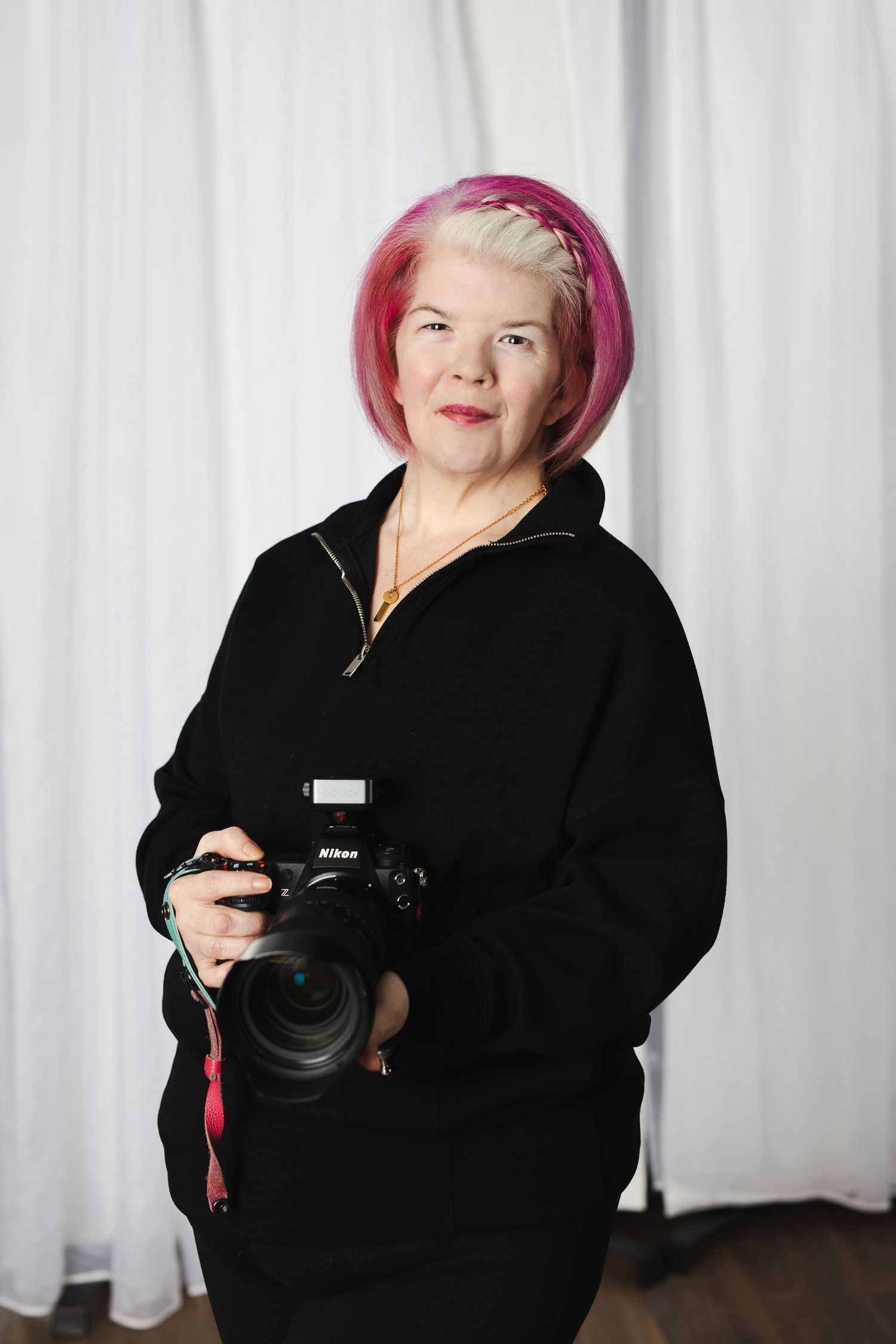 Professional photographer wearing a black hoodie and pink hair holds a Nikon camera against white curtain backdrop.