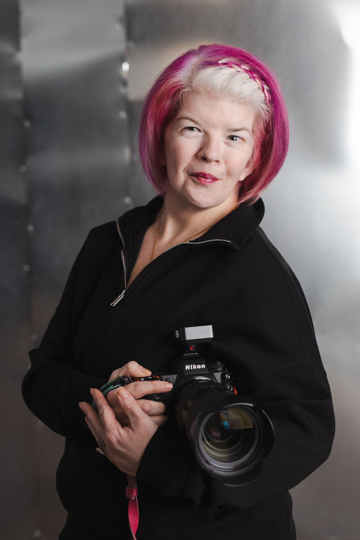 Professional photographer with bright pink hair wearing a black coat holds a Nikon DSLR camera in a studio setting.