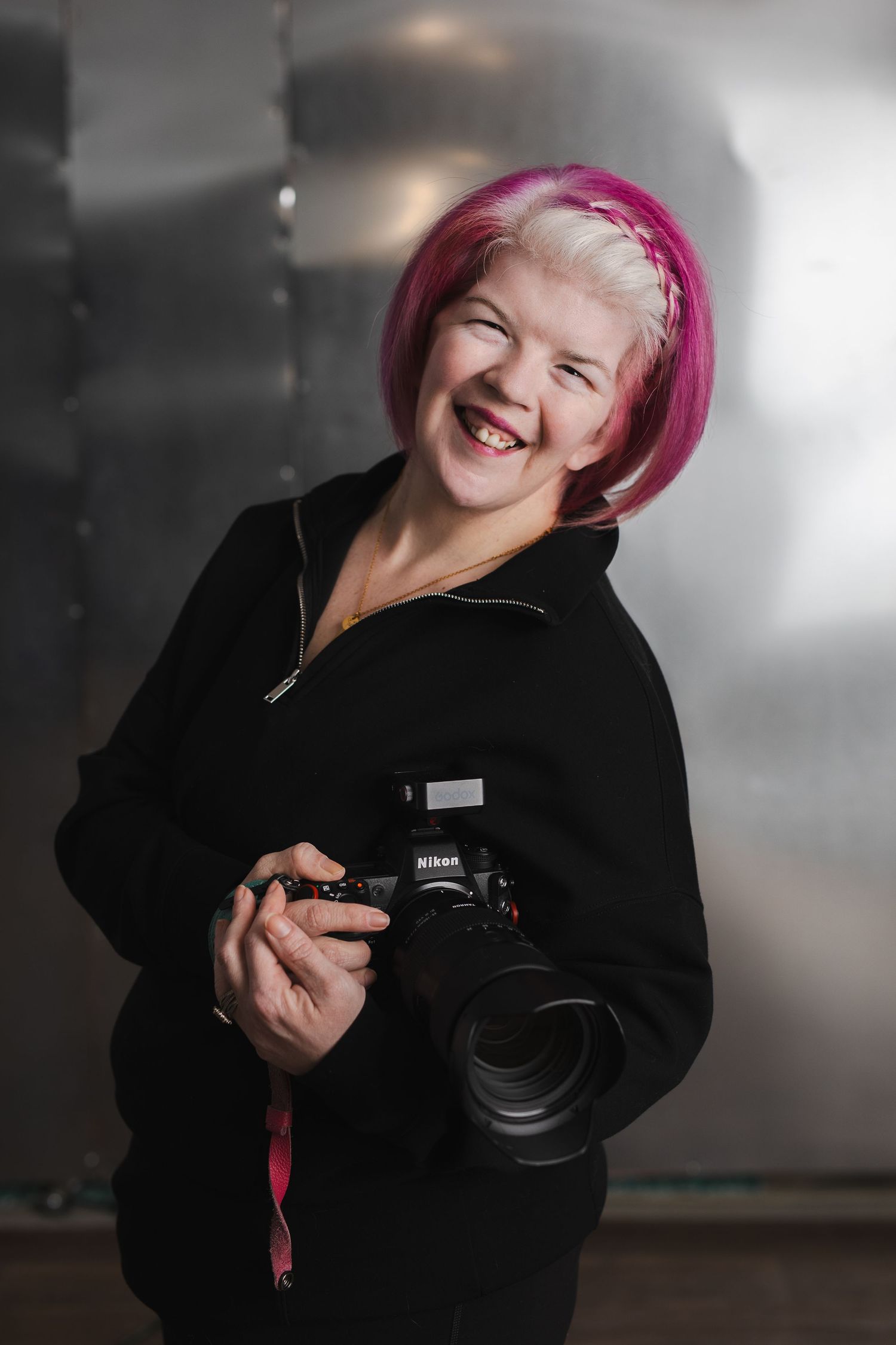 Professional photographer in black jacket with pink hair laughs joyfully while holding camera in studio setting.