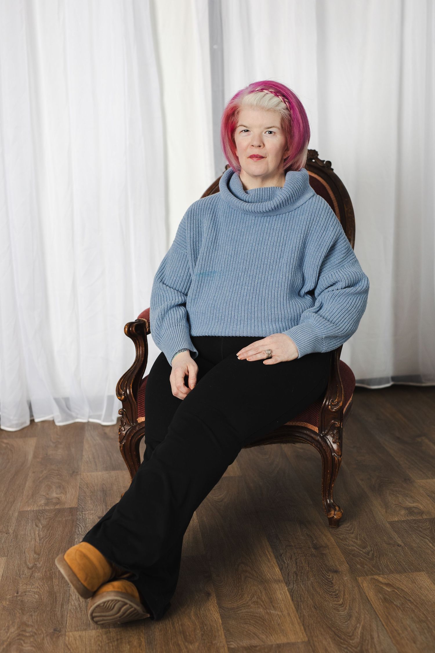 Person with pink hair wearing a blue sweater and black pants sits in a wooden chair on hardwood floor.