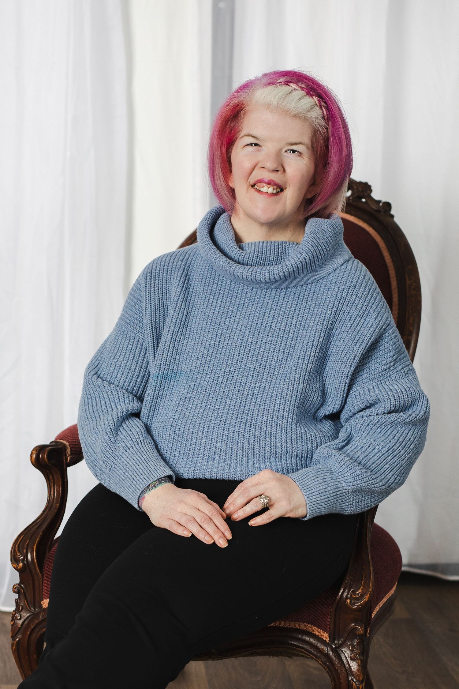 A person wearing a blue sweater and black pants sits in an antique wooden chair, laughing joyfully.