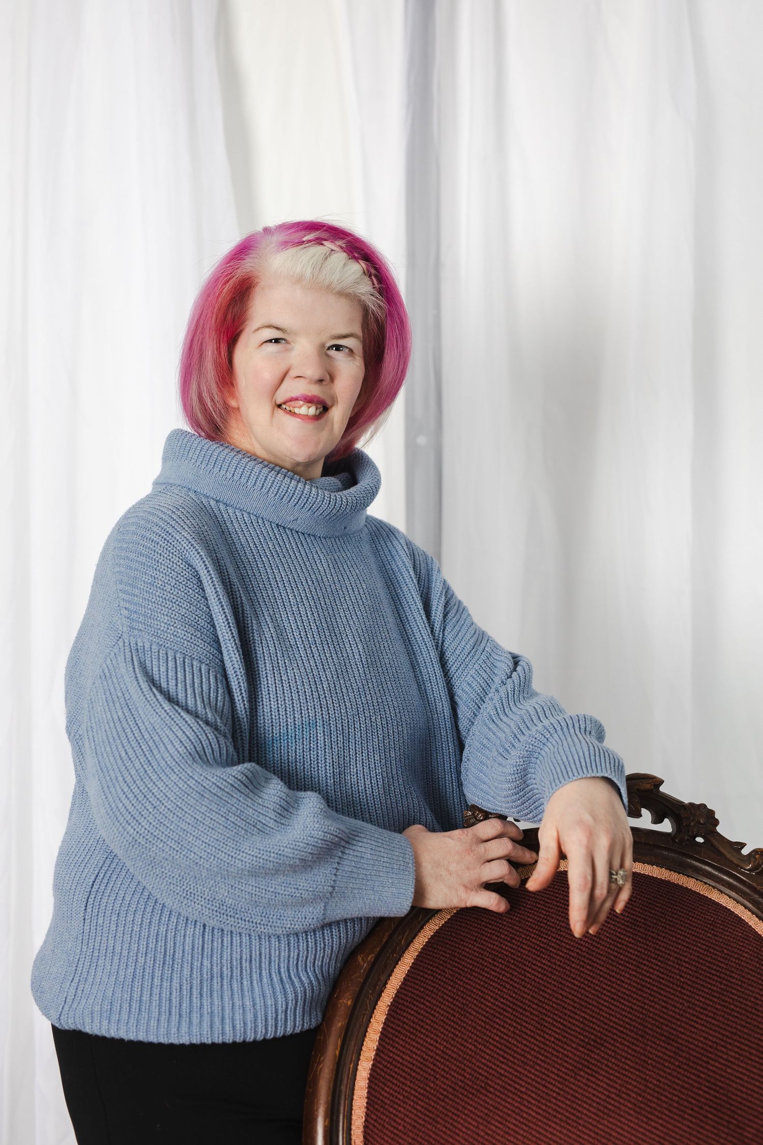 A person with pink hair wearing a blue sweater poses playfully while leaning on a brown chair.