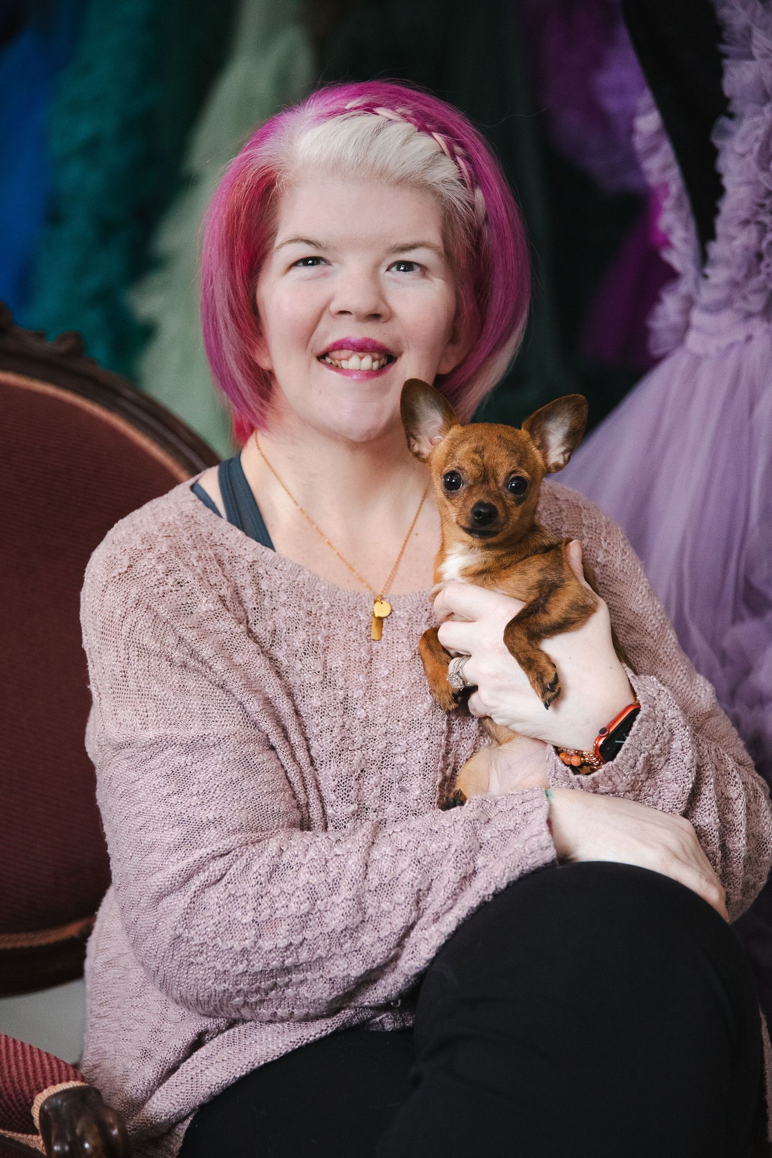 A small brown Chihuahua puppy being held by someone wearing a cozy pink sweater against a colorful backdrop.