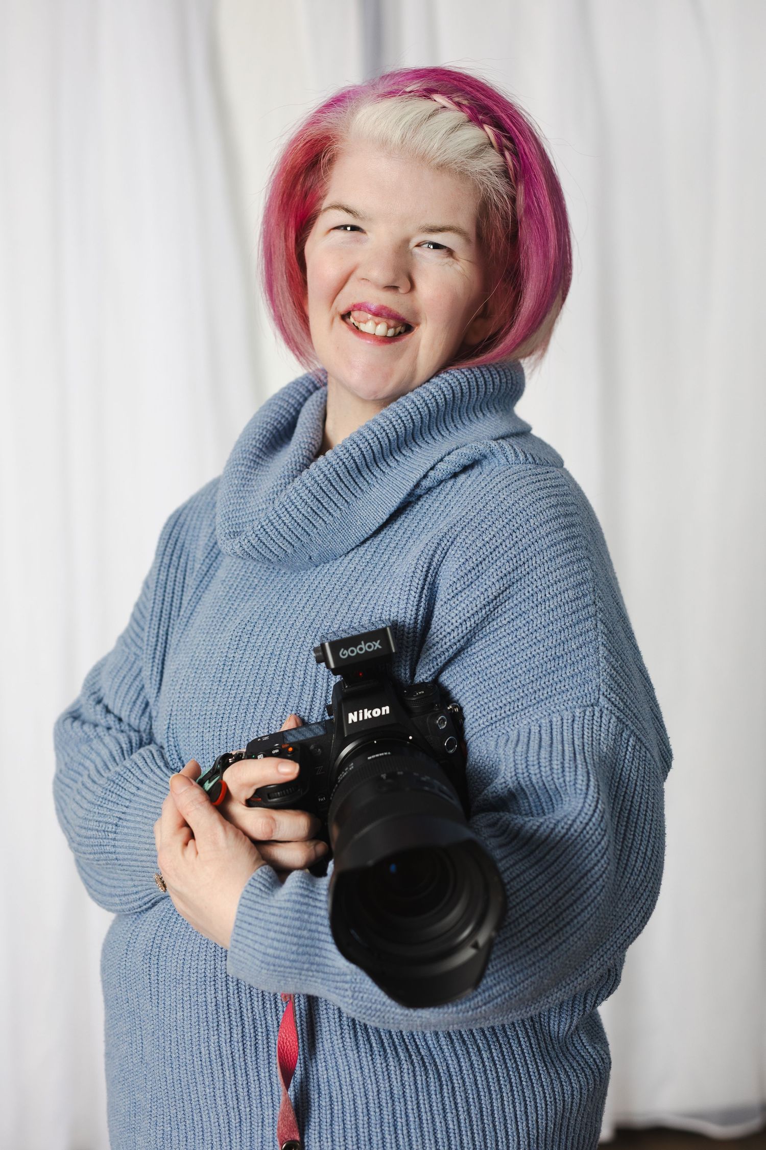 Photographer wearing a cozy gray sweater holds a camera while posing playfully against a white curtain background.
