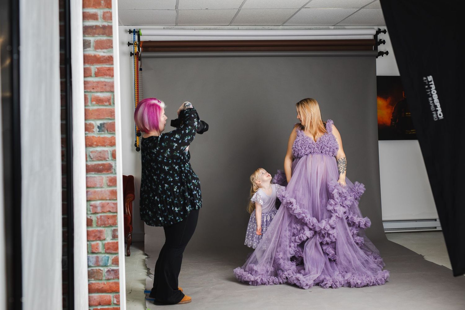 Behind the scenes photo shoot of a model wearing a flowing purple gown in a studio with brick walls.