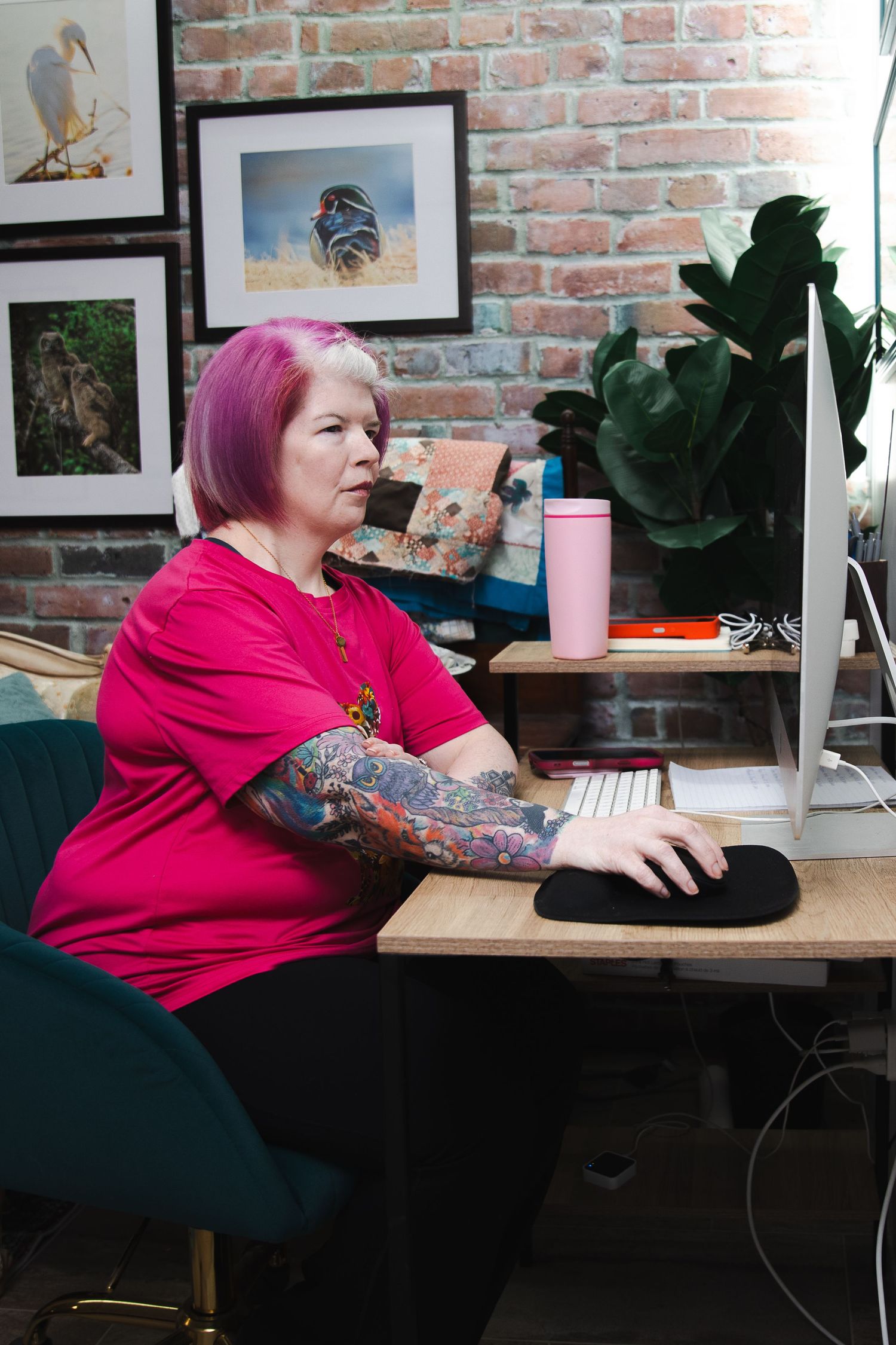 A person with colorful tattoos and pink hair sits at a desk with houseplants and artwork in a brick-walled room.