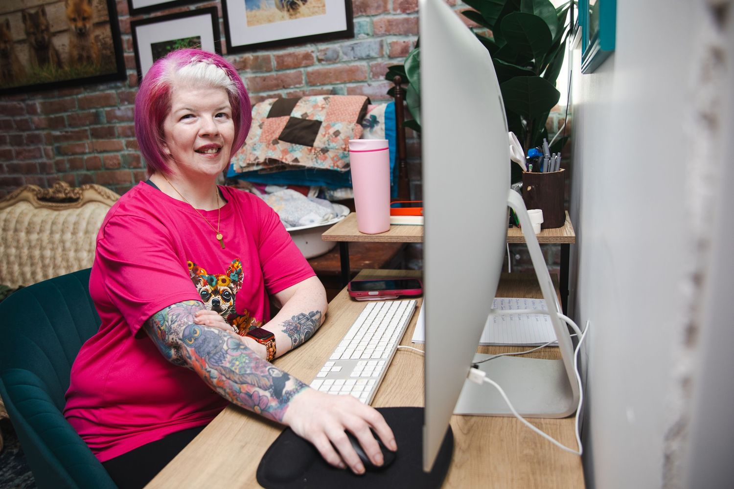 A person in a bright pink shirt works at a desk with computers and office equipment in a brick-walled workspace.