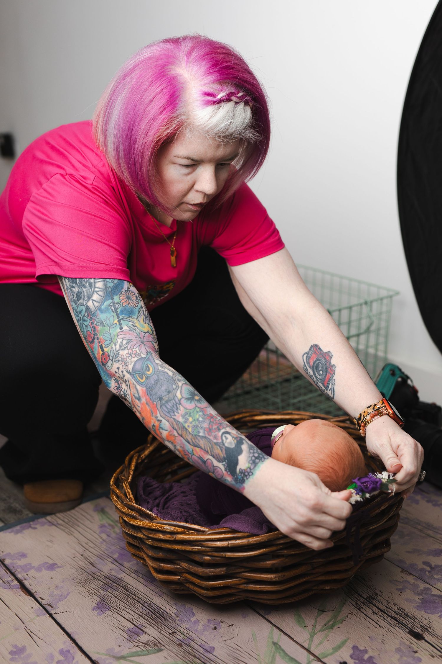 A person with pink hair and tattooed arms repairs a brown woven basket on a rustic wooden surface.