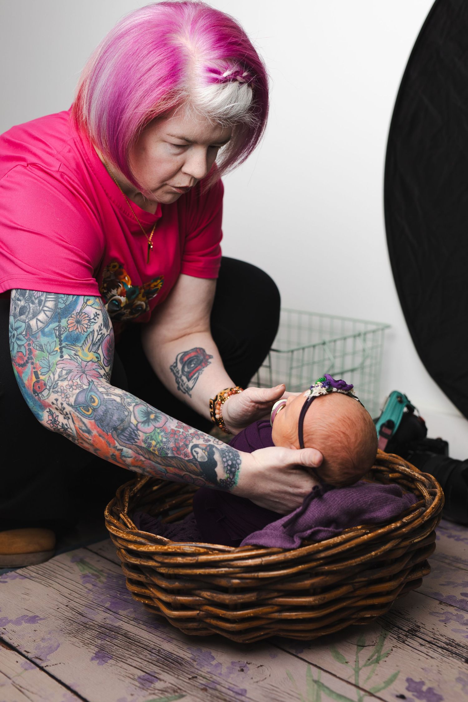 A person with pink hair and tattooed sleeves arranges items in a woven basket on a rustic wooden surface.