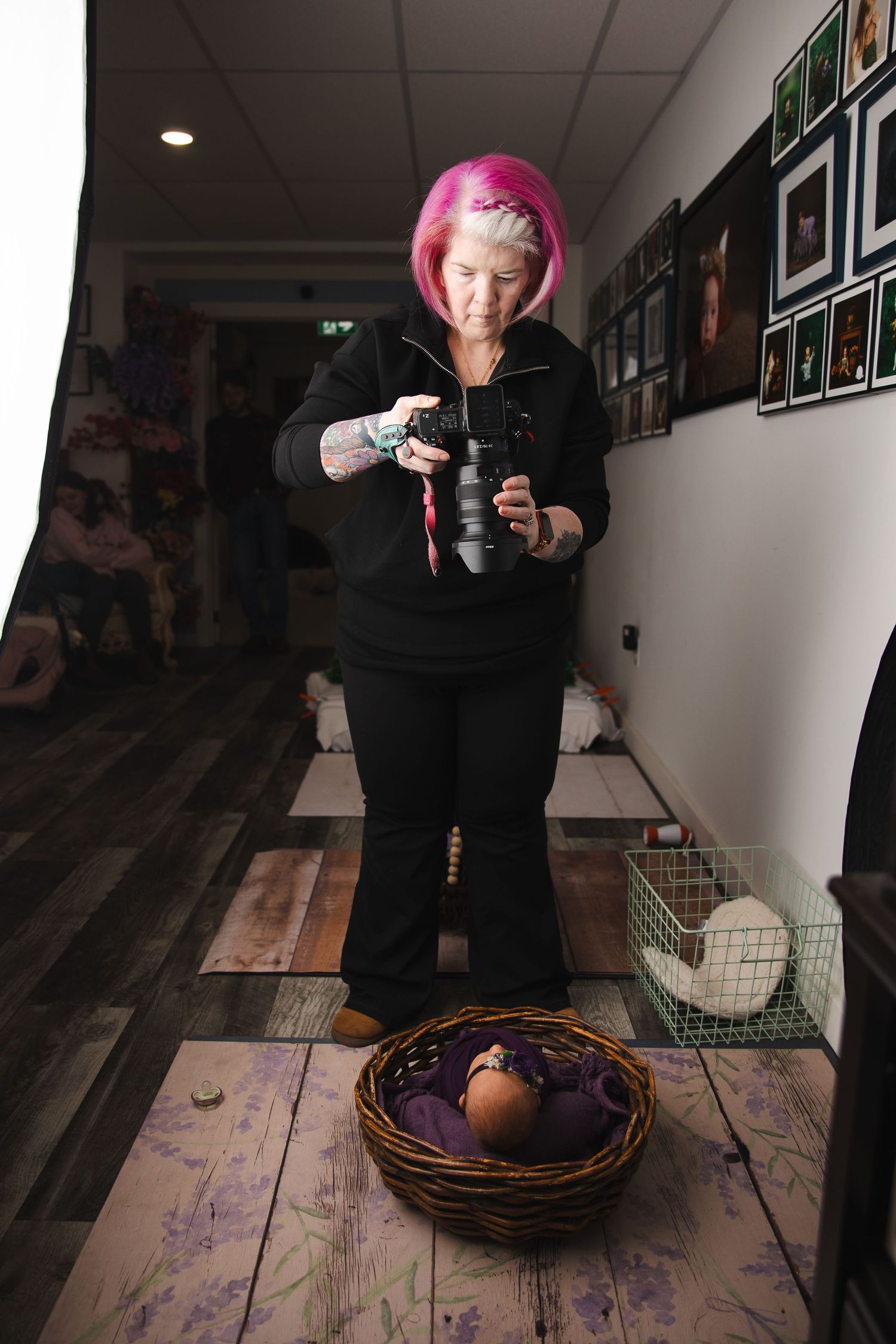 A person with pink hair in black clothing stands with a camera while a decorative basket sits on a wooden floor.