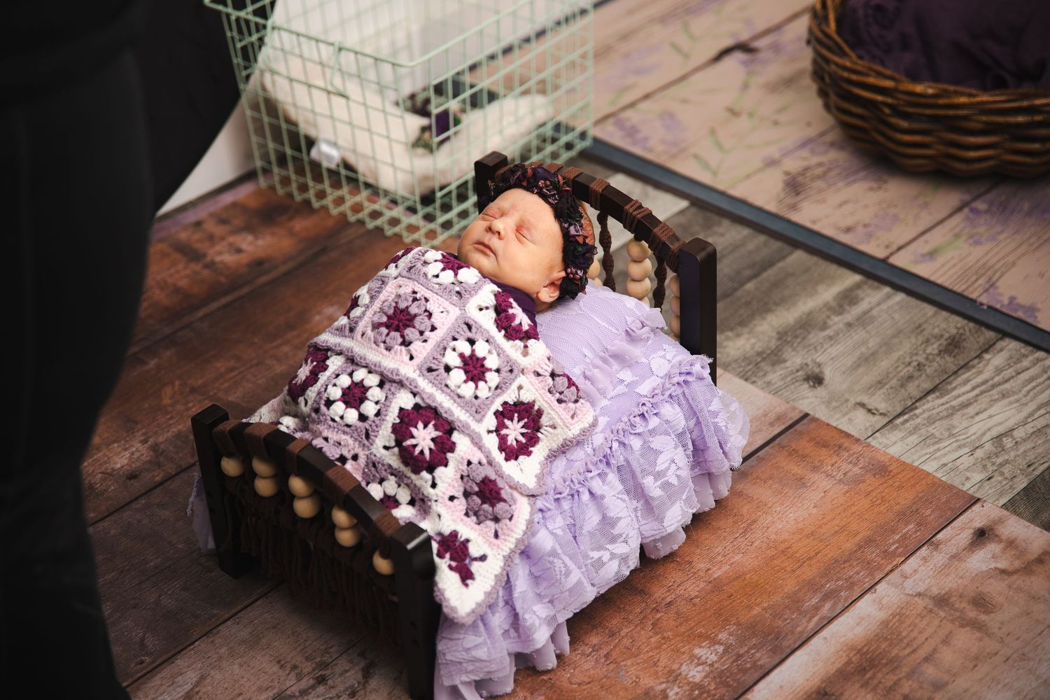 A newborn baby wrapped in purple floral lace fabric lies sleeping on a rustic wooden surface.
