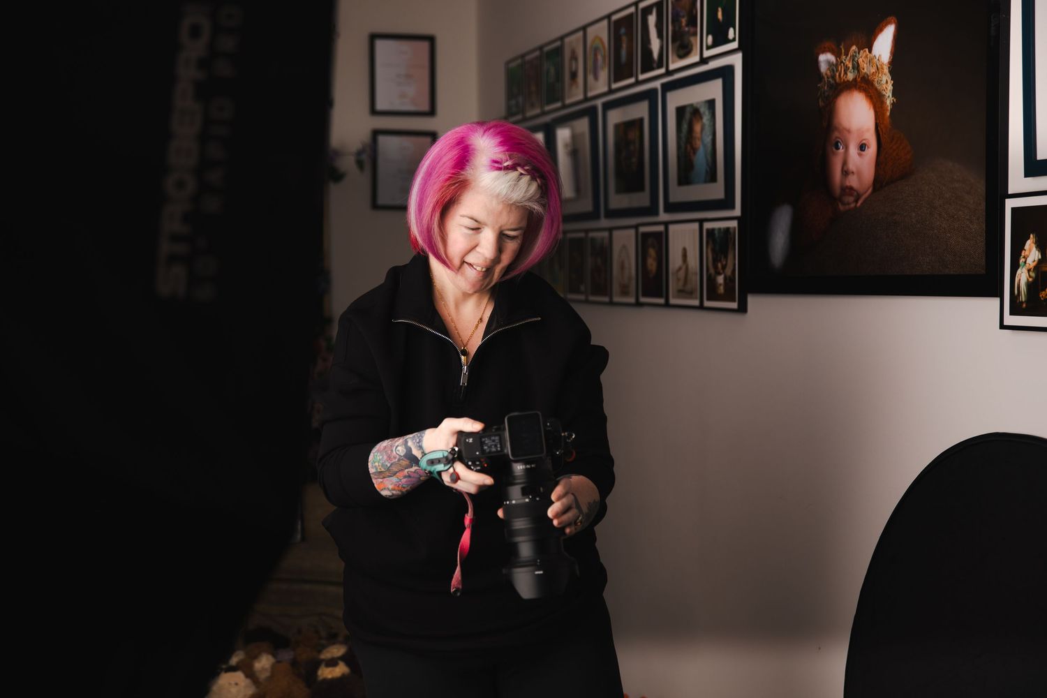 A photographer with bright pink hair wearing black clothing holds a camera in a studio with framed photos on the wall.